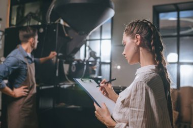 selective focus of woman checking colleagues work during coffee roasting process clipart