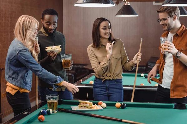 company of multiethnic friends eating and drinking beside pool table at bar