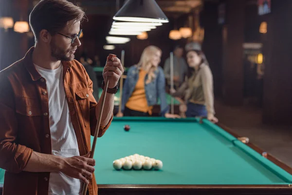young man chalking cue beside pool table at bar with friends