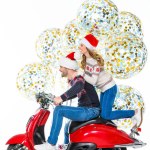 Cheerful couple in santa hats riding red scooter, big christmas balloons with confetti on background, isolated on white