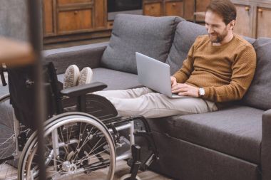 smiling disabled man putting legs on wheelchair while using laptop on sofa at home clipart