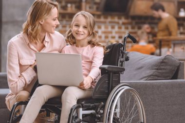 happy mother looking at cute little daughter sitting in wheelchair and using laptop at home