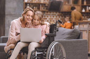 Anne ve küçük kızı evde birlikte dizüstü kullanarak tekerlekli sandalyede gülümseyen 