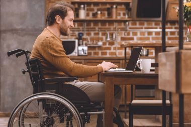 side view of smiling disabled man in wheelchair using laptop at home clipart