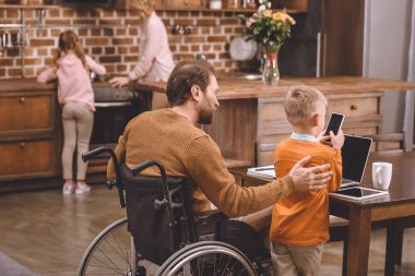Arkadan görünüş Engelli babasının tekerlekli sandalye ve küçük oğlu smartphone ile dizüstü birlikte evde kullanma