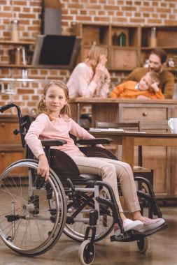 cute little girl in wheelchair looking at camera while other members of family standing behind at home clipart