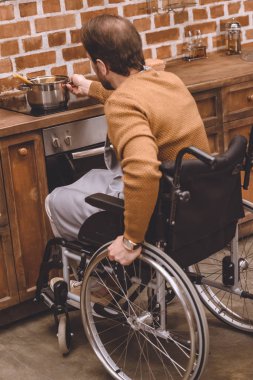 back view of disabled man in wheelchair holding pan while cooking at home