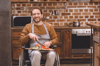 disabled man in wheelchair cutting vegetables and smiling at camera at home