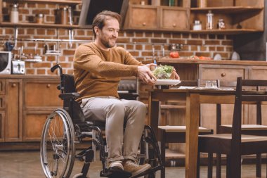 happy disabled man in wheelchair holding glass bowl of vegetable salad at home  clipart