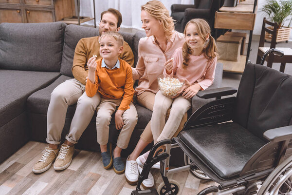 happy family sitting on sofa and eating popcorn, wheelchair standing near by