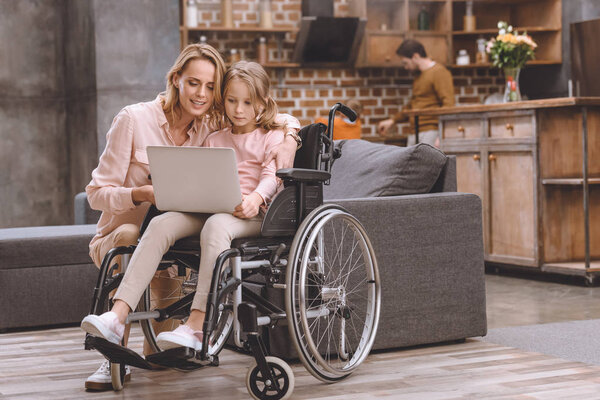 smiling mother and little daughter in wheelchair using laptop together while father and son standing behind at home 