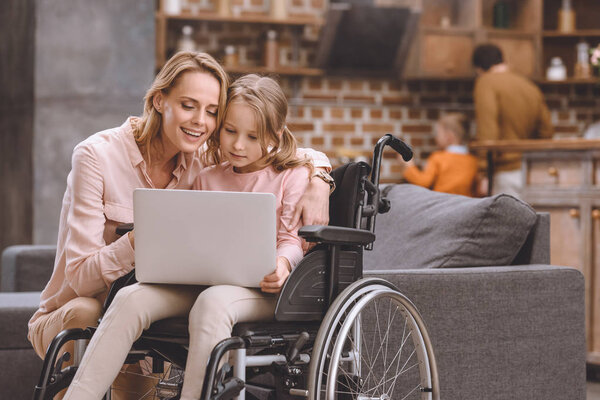 smiling mother and little daughter in wheelchair using laptop together at home 