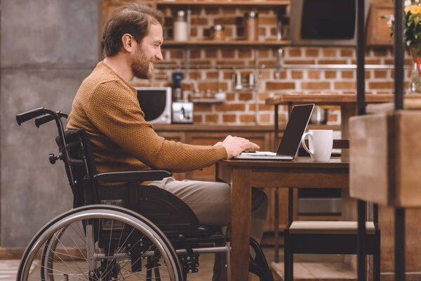 side view of smiling disabled man in wheelchair using laptop at home