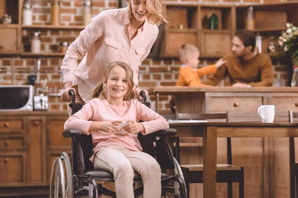 Madre Guardando Sorridente Figlia Disabile Seduta Sedia Rotelle Utilizzando Smartphone — Foto Stock