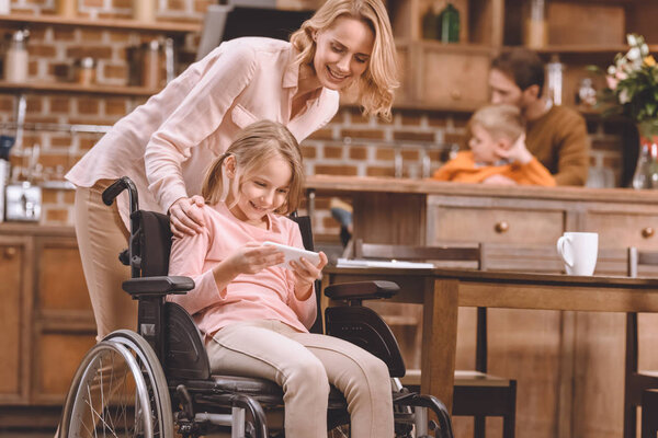 happy mother looking at smiling disabled daughter sitting in wheelchair and using smartphone at home 