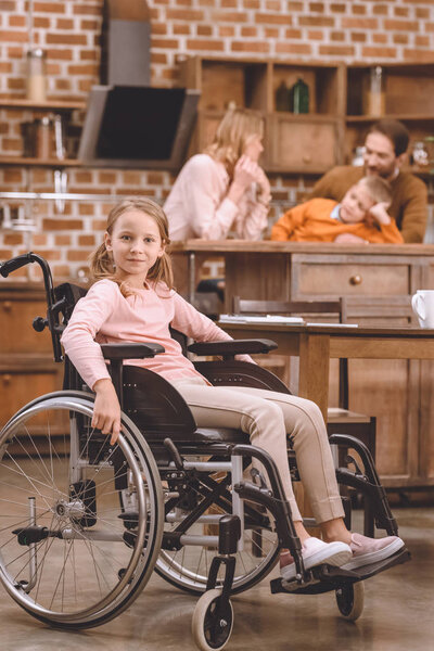cute little girl in wheelchair looking at camera while other members of family standing behind at home