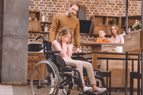 father looking at little daughter sitting in wheelchair and closing eyes with palms at home
