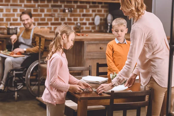 Hermanos Con Madre Sirviendo Mesa Para Cena Mientras Padre Discapacitado —  Fotos de Stock