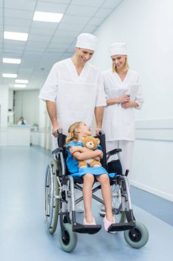 doctors and little patient with teddy bear in wheelchair in hospital clipart