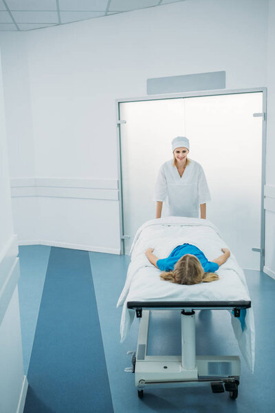 smiling doctor moving preschooler patient on medical stretcher in corridor 