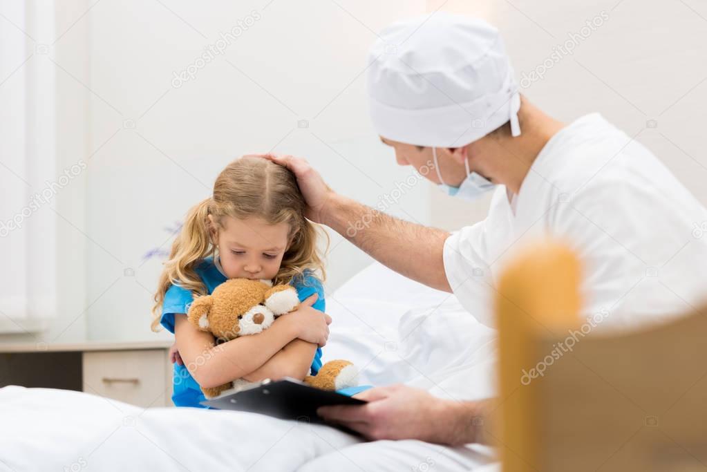 doctor palming head of worried kid with soft toy