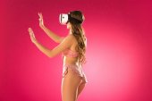 side view of seductive woman in underwear touching something with virtual reality headset isolated on pink