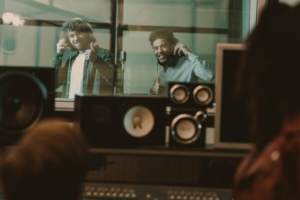 young handsome musicians recording song behind glass at studio and showing thumbs up