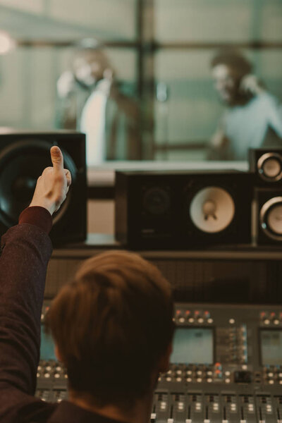 sound producer showing thumb up to singers behind glass at recording studio
