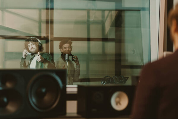 young happy musicians recording song behind glass at studio and showing thumbs up