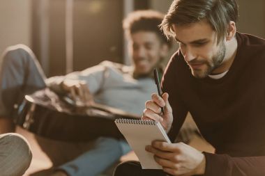 handsome young man writing lyrics in notepad while his friend lying on floor with guitar clipart