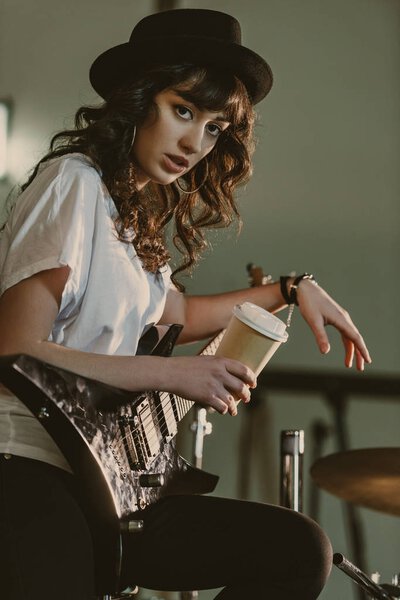 beautiful female musician with electric guitar and cup of coffee to go looking at camera