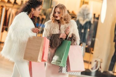fashionable multiethnic women in fur coats holding paper bags and shopping together in mall 