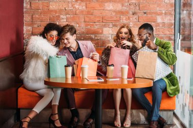 smiling trendy multiethnic young people looking into paper bags while drinking coffee together   clipart