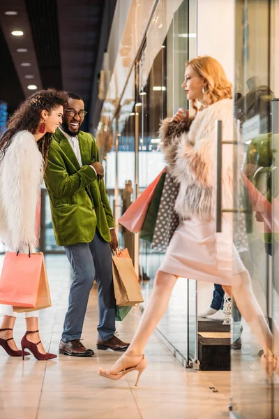 时尚女人的侧面看着购物袋和步行 而微笑的非洲裔美国夫妇站在商场后面 — 图库照片