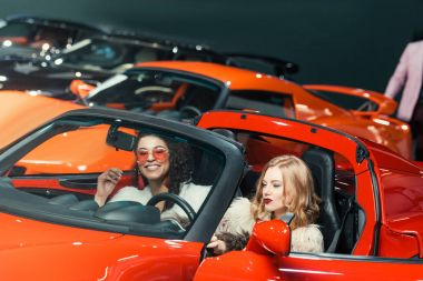 fashionable smiling multiethnic women sitting in luxury red car clipart