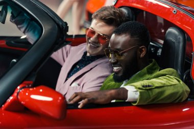 happy stylish multicultural men sitting in new car at showroom clipart
