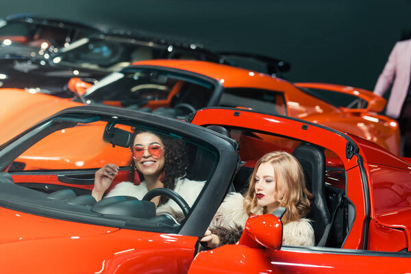 fashionable smiling multiethnic women sitting in luxury red car
