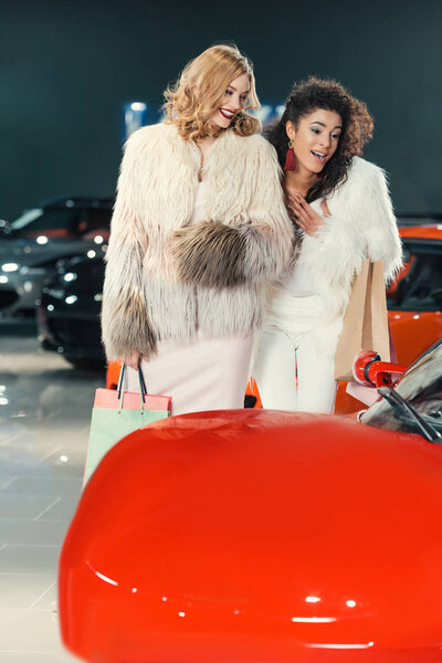 young women in fur coats looking at car in showroom