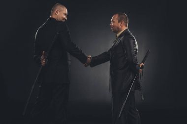 side view of businessmen shaking hands with katanas behind back isolated on black clipart