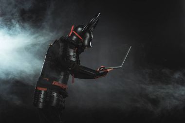 side view of of samurai using laptop on dark background with smoke clipart