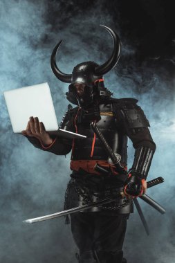 samurai in traditional armor holding laptop on dark background with smoke clipart