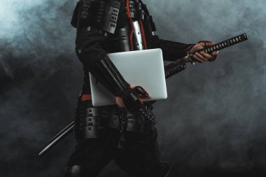 cropped shot of samurai in traditional armor with laptop taking out sword on dark background with smoke clipart