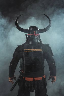 back view of samurai in traditional armor and horned helmet on dark background with smoke clipart