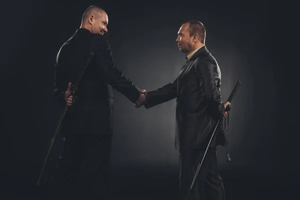 stock image side view of businessmen shaking hands with katanas behind back isolated on black