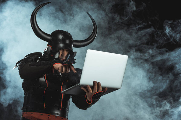 armored samurai using laptop on dark background with smoke