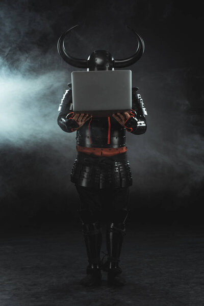 бронированный самурай держит ноутбук на темном фоне с дымом
