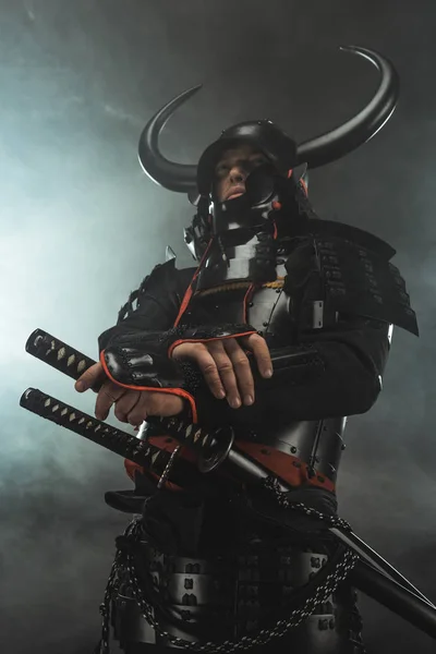 bottom view of samurai in traditional armor with swords on dark background with smoke