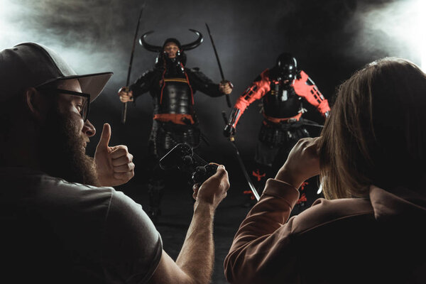 rear view of man and woman playing samurai fighting in real life with gamepads on black