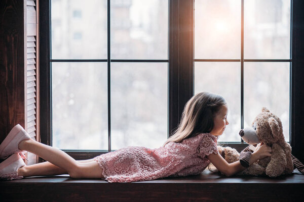 side view of little child playing with teddy bear while lying on windowsill