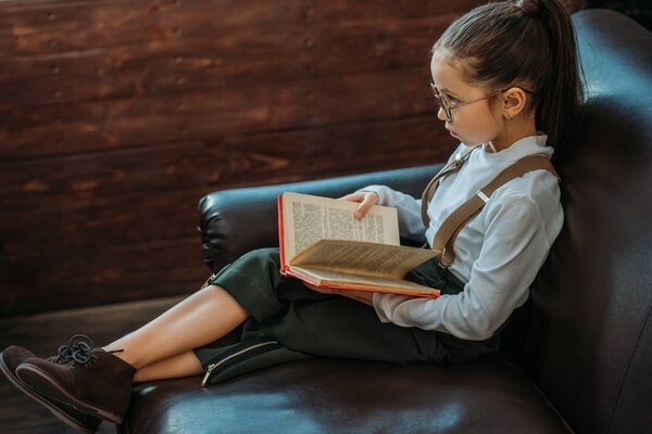 serious little child with book sitting on couch at home